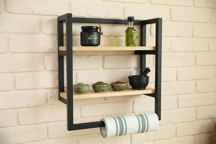 A black metal and light wood three-tier wall-mounted spice rack with a paper towel holder, styled with kitchen items against a white brick wall.