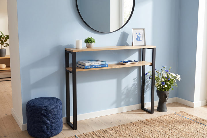 A two-tier console table with light wood shelves and black metal legs, styled in a bright room with a blue wall, round mirror, and decorative items.