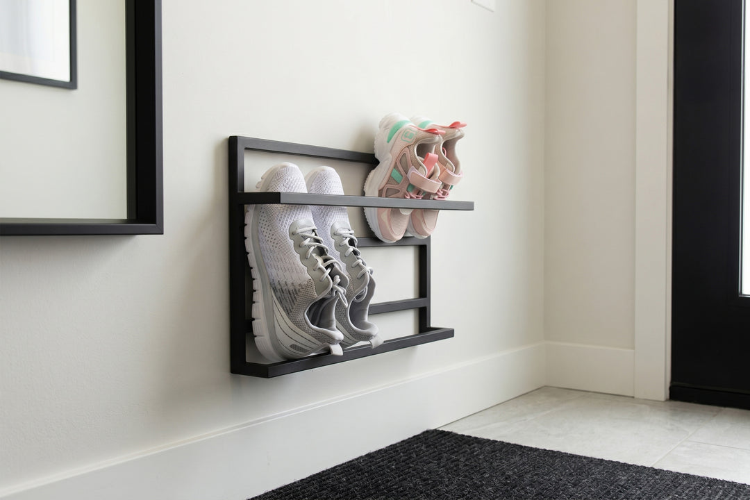 A black metal wall-mounted shoe rack holding a pair of white sneakers and a pair of pastel sneakers, mounted on a white wall in a clean entryway setting.