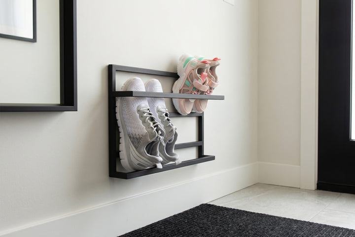 A black metal wall-mounted shoe rack holding a pair of white sneakers and a pair of pastel sneakers, mounted on a white wall in a clean entryway setting.