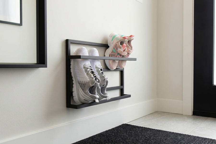 A black metal wall-mounted shoe rack holding a pair of white sneakers and a pair of pastel sneakers, mounted on a white wall in a clean entryway setting.